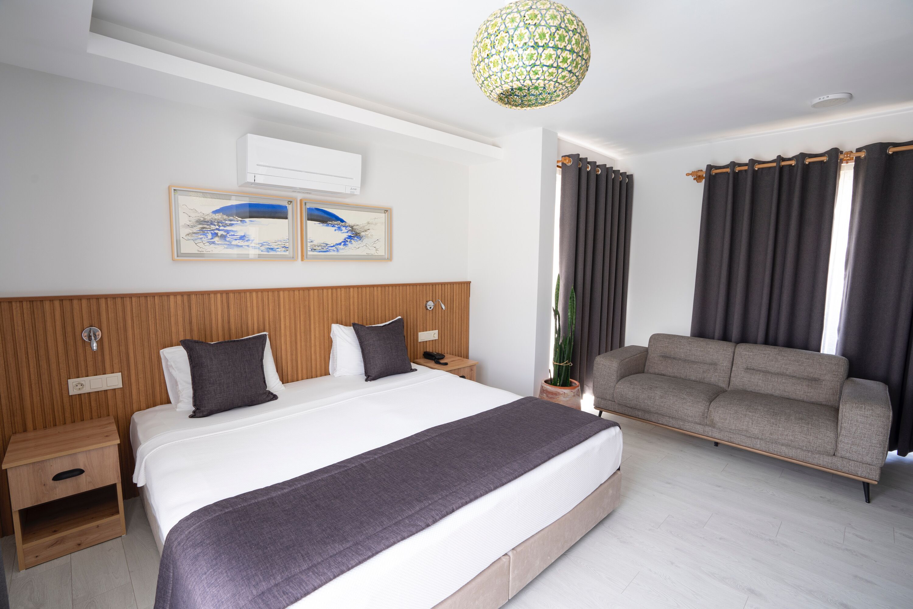A hotel room with a large bed, a sofa, a nightstand, a potted plant, and artwork on the wall.
