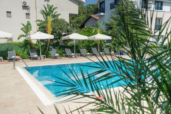 Piscina com espreguiçadeiras e guarda-sóis, rodeada por vegetação e edifícios.