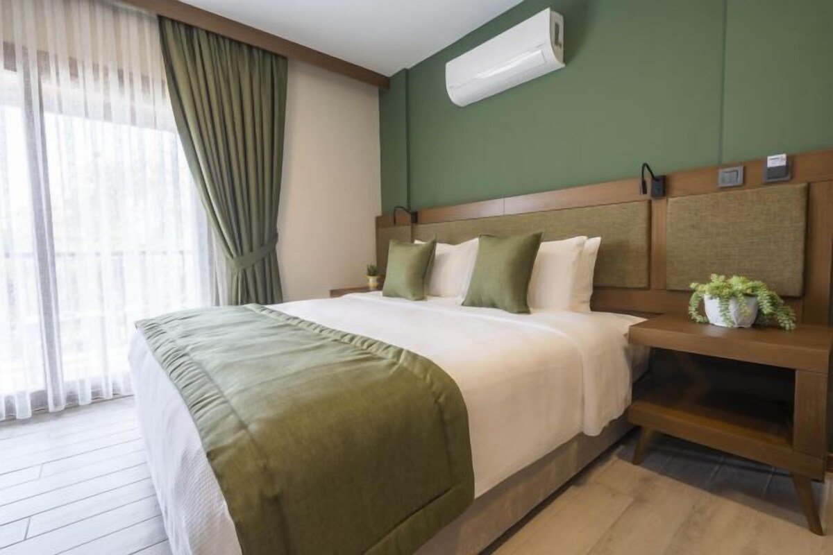 A hotel room with a bed, a nightstand, a potted plant, and a window with curtains.