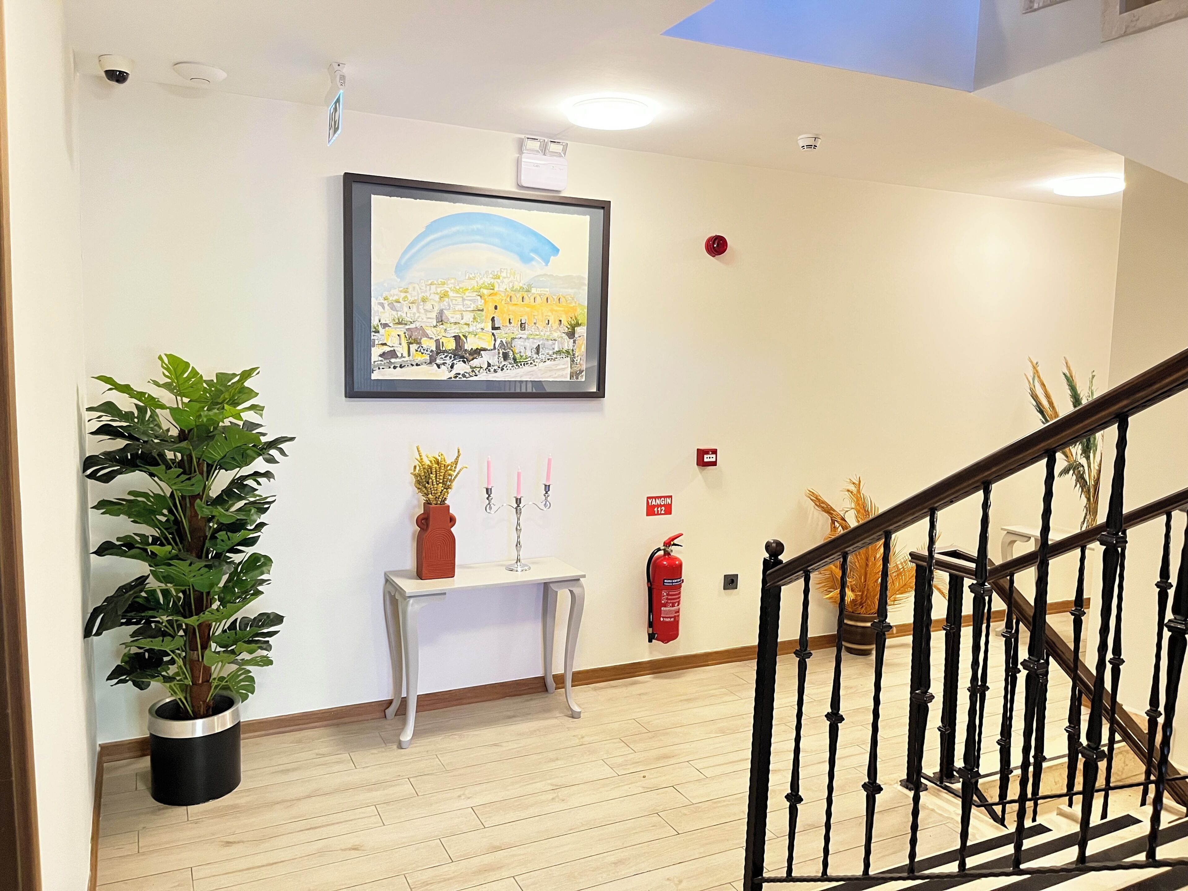 A hallway with a framed painting, a potted plant, a small table with a vase and candelabra, a fire extinguisher, and a staircase.