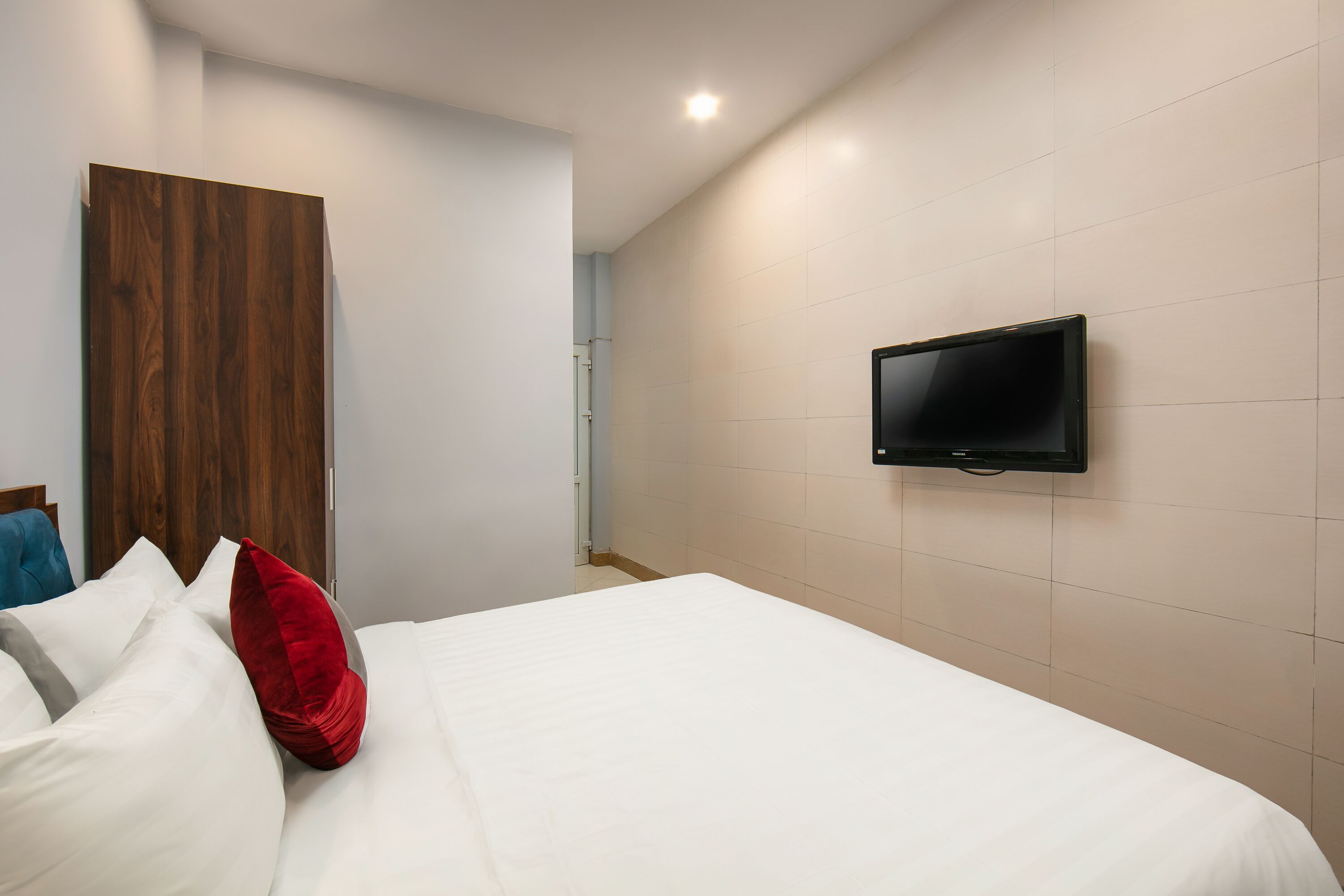 Une chambre d’hôtel avec un lit, du linge de lit blanc, un oreiller rouge, une armoire en bois et un téléviseur fixé au mur.