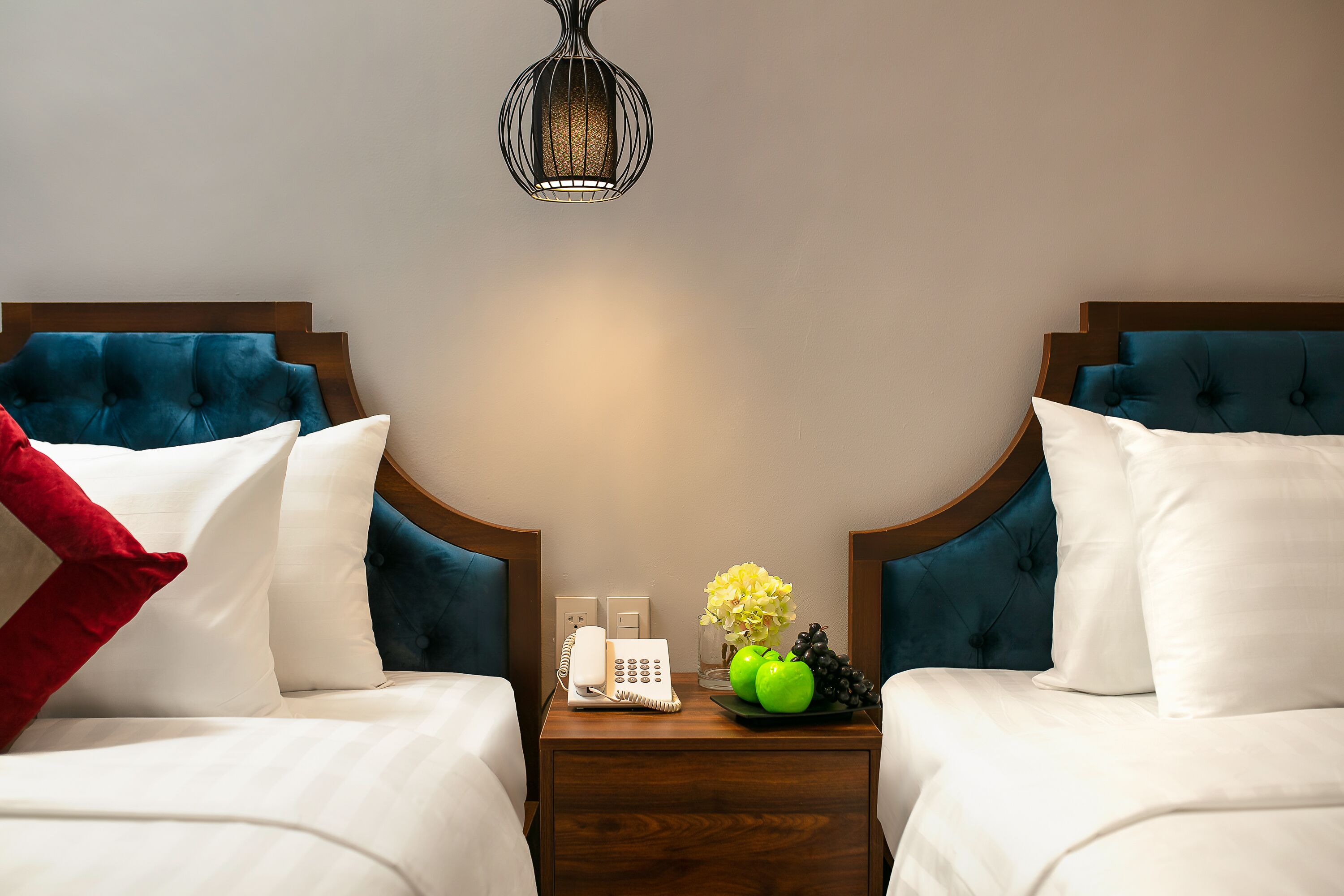 A hotel room with two beds, a nightstand with a phone and a vase of flowers, and a hanging light fixture.