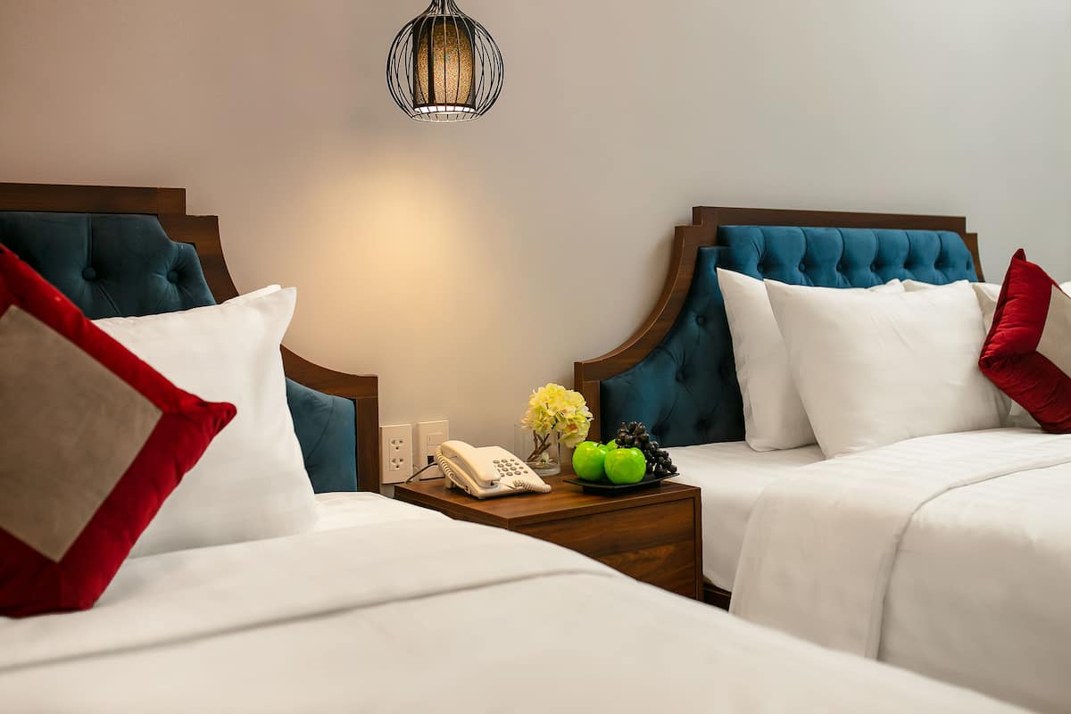 Une chambre d’hôtel avec deux lits, chacun doté d’une tête de lit en bois, de draps blancs et d’oreillers rouges et blancs. On y trouve une table de chevet avec un téléphone, un vase de fleurs et une assiette contenant des pommes et des raisins.