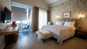 A bedroom with a stone wall, a bed with a striped bedspread, a bedside table with a lamp, a chair, and a desk with a television. - Zena Hotel Alaçatı Adults Only (Cesme)