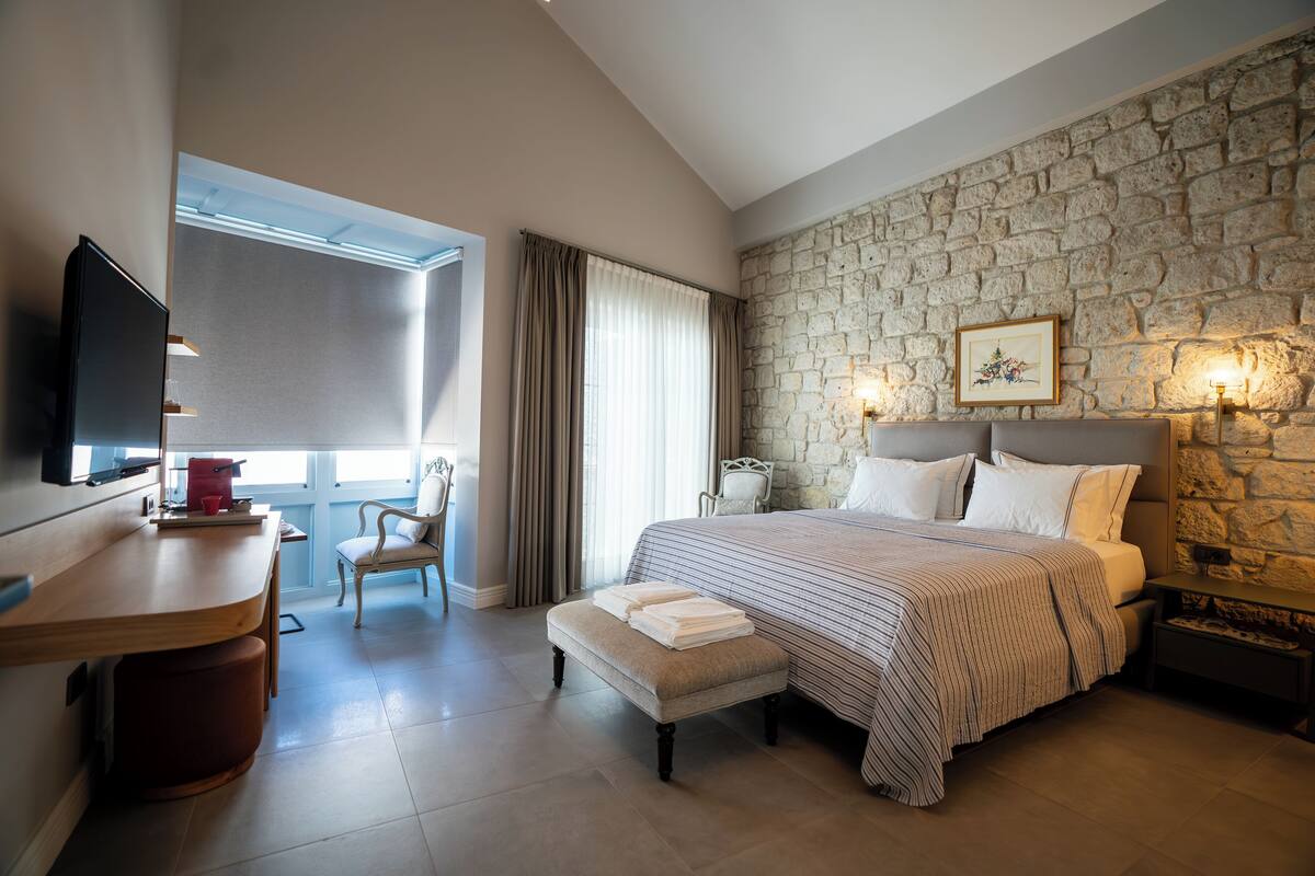 A bedroom with a stone wall, a bed with a striped bedspread, a bedside table with a lamp, a chair, and a desk with a television.