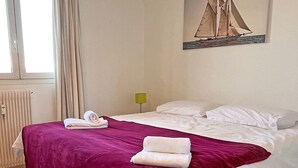 Een net opgemaakt bed met een paarse deken en witte kussens, een schilderij van zeilboten aan de muur en een raam met gordijnen.