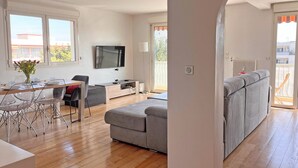 Een moderne woonkamer met een grijze bank, een eettafel met stoelen, een aan de muur bevestigde televisie en een balkon met uitzicht.