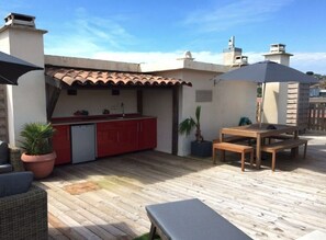 Un patio con terraza de madera, una zona de cocina roja, una mesa de madera con bancos y una planta en maceta.