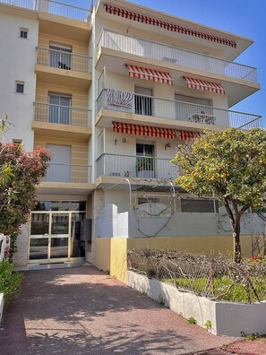 Un edificio residencial de varios pisos con balcones, entrada protegida por un portón y un pequeño jardín.