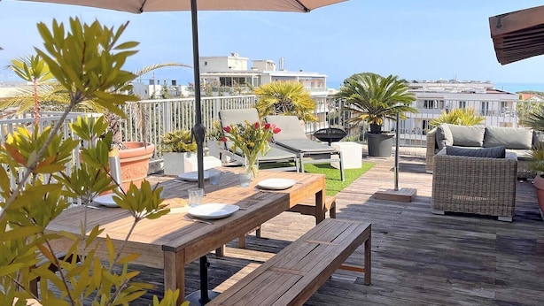 Een houten terras met een tafel, stoelen, een grote parasol en potplanten.
