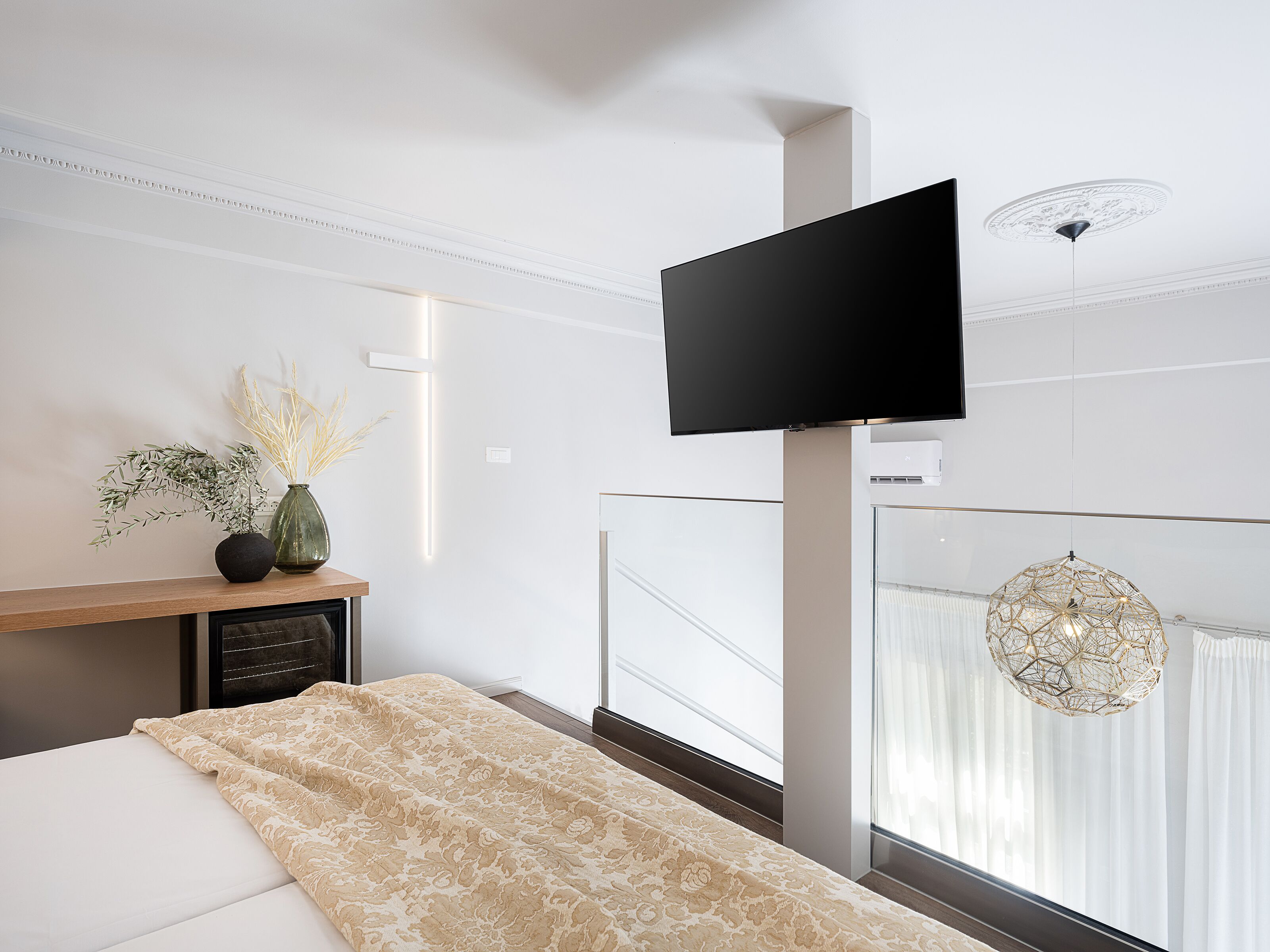 A modern bedroom with a bed, a flat-screen TV mounted on the wall, a bedside table with a vase, and a view of the city through large windows.