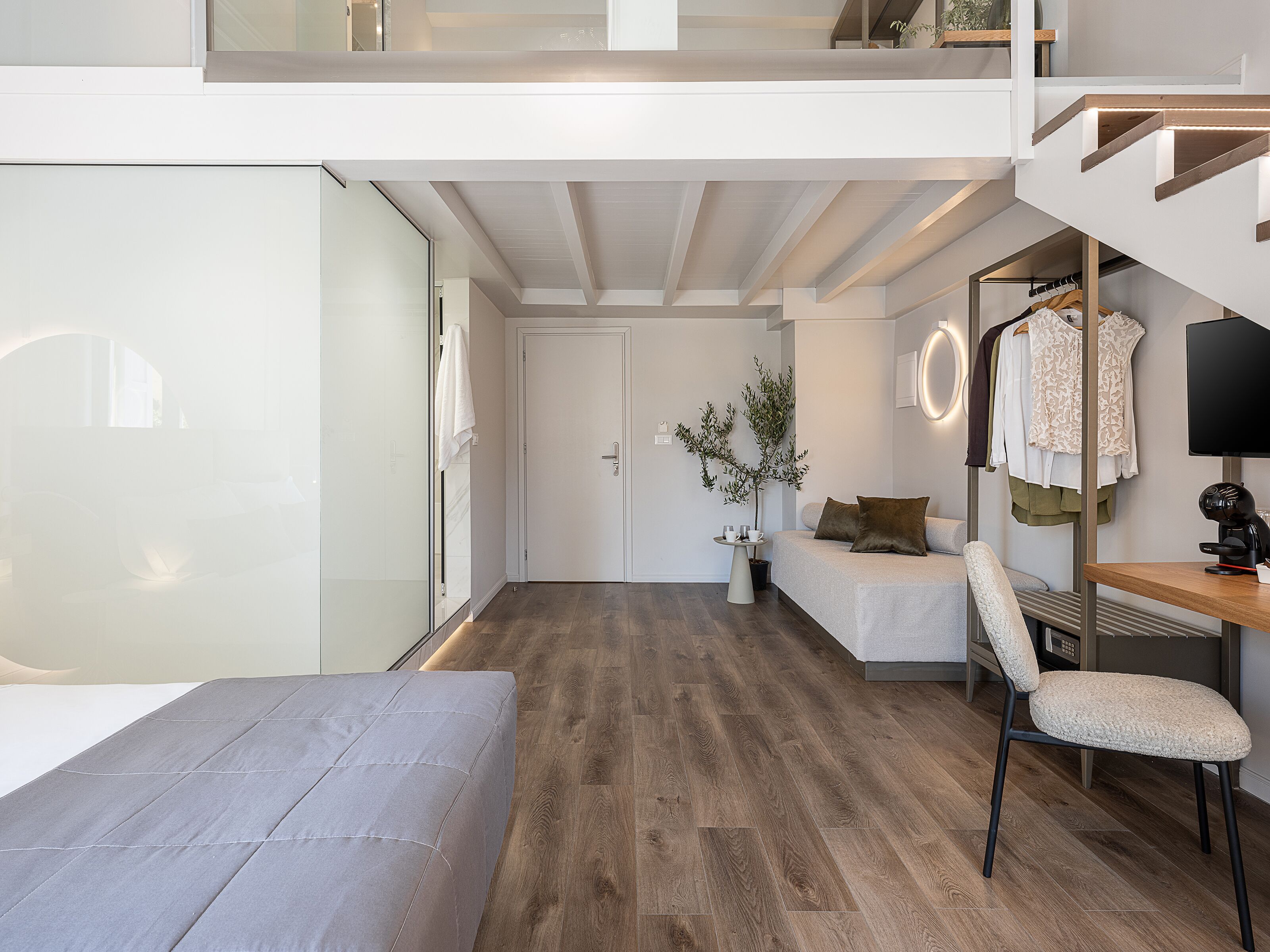 Et moderne hotelværelse med trægulv, seng, skrivebord med stol, knagerække og trappe.
