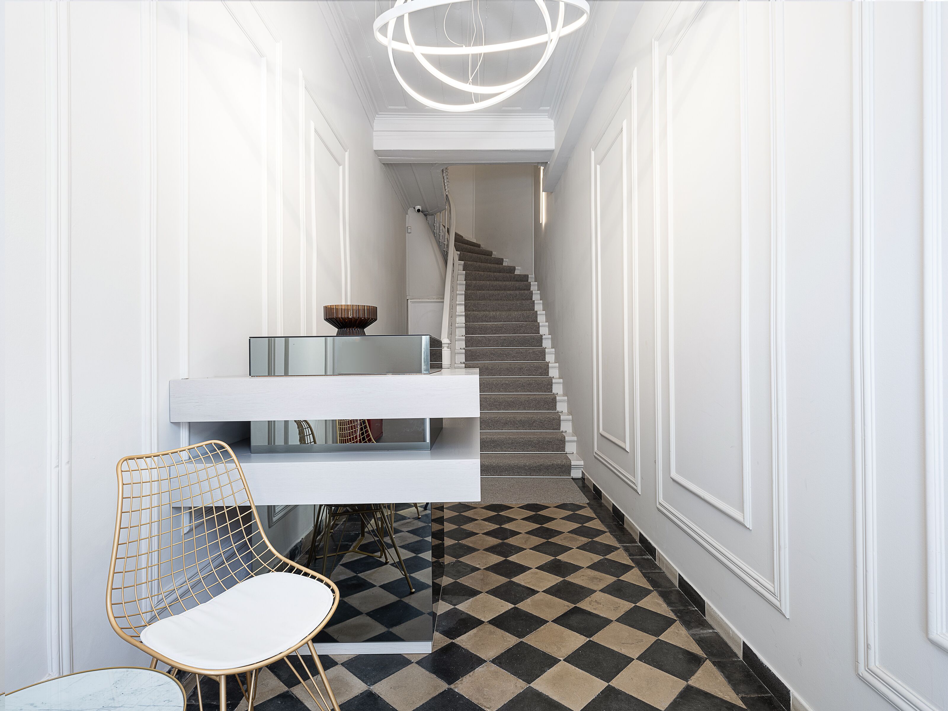 A hallway with a checkered floor, a staircase, a white wall, and a chair.