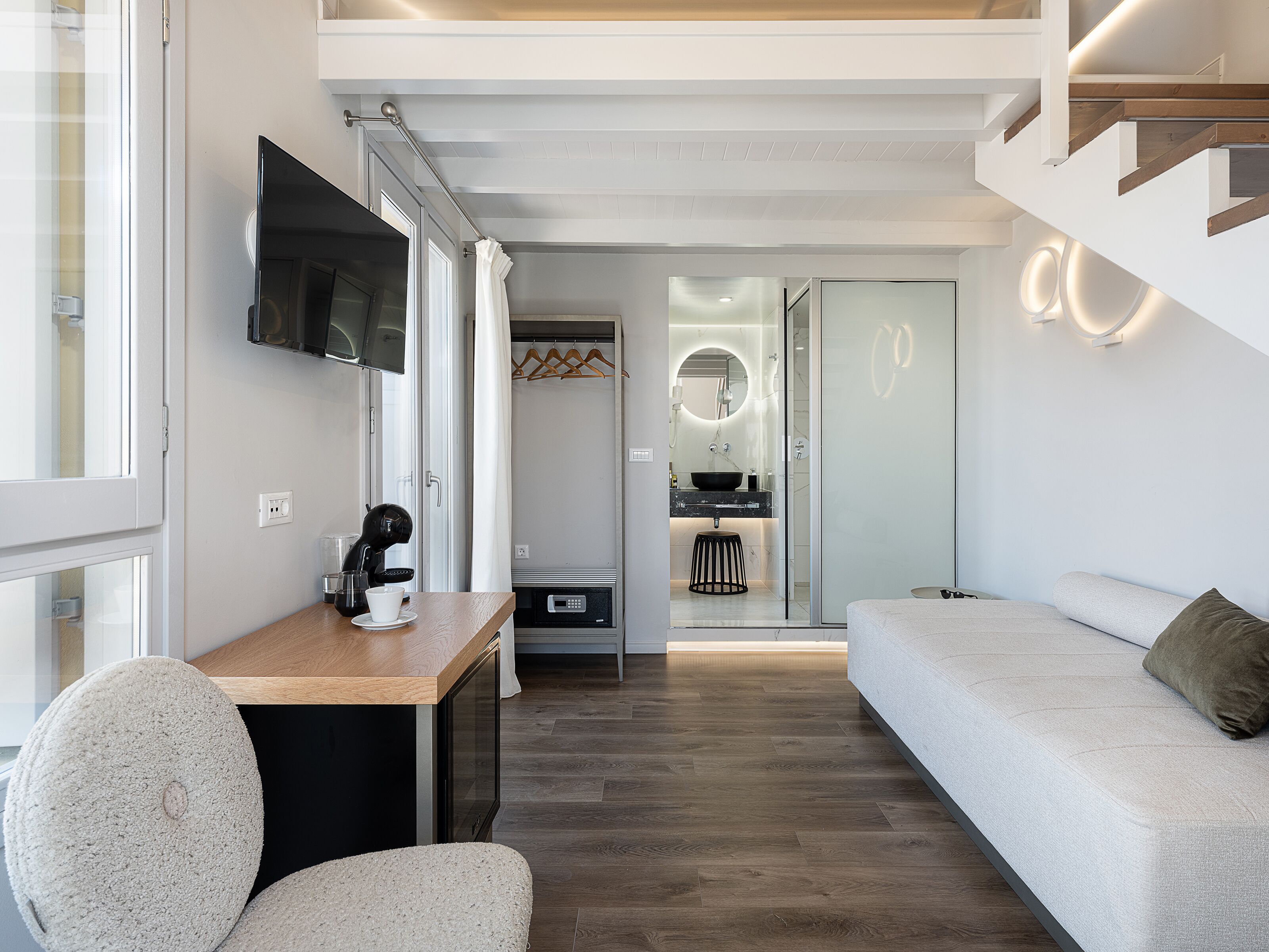 Una habitación moderna y bien iluminada, con piso de madera, un sofá, una mesa de centro, un televisor y un baño visible a través de una puerta de vidrio.