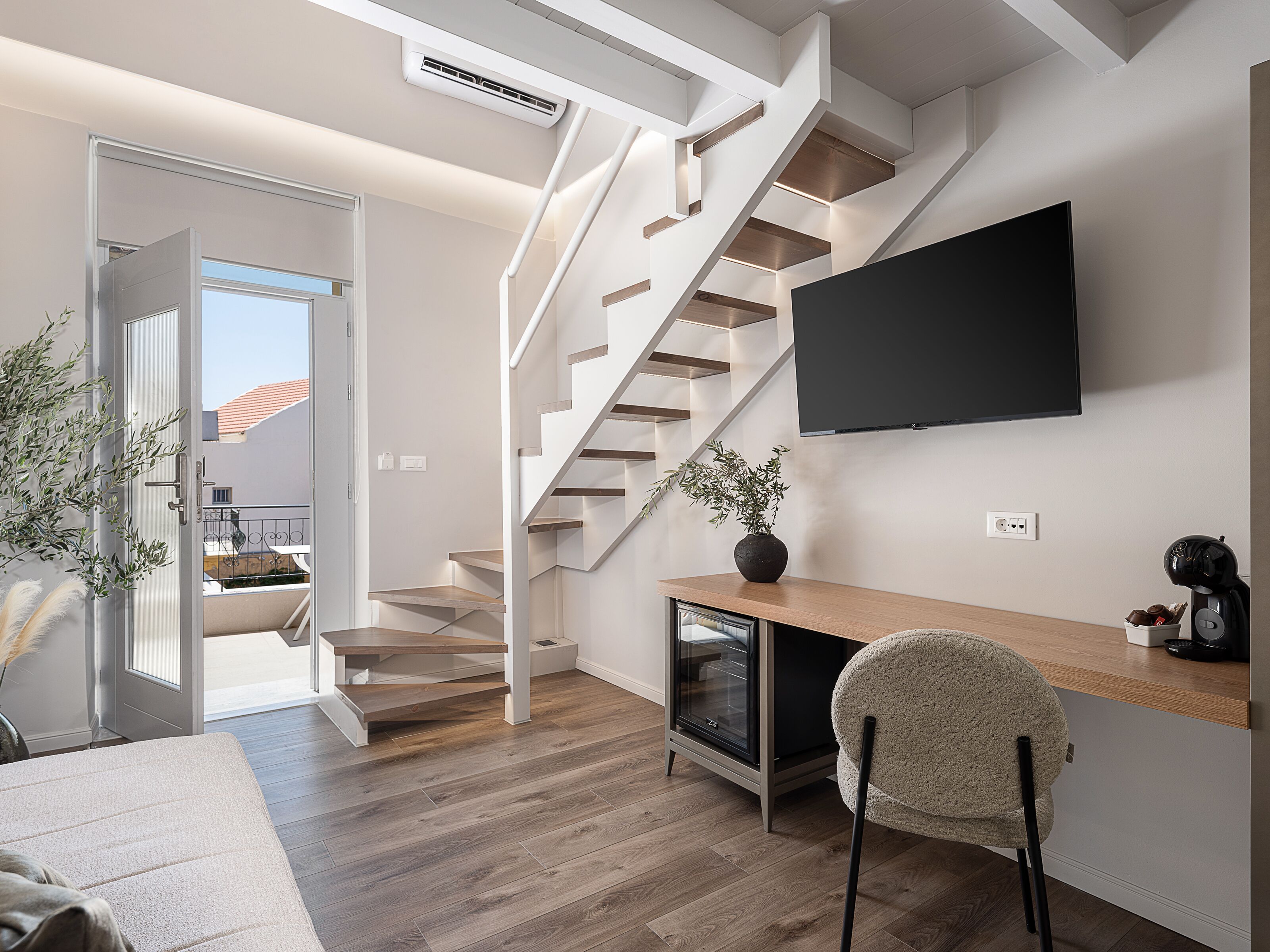 A modern living room with a wooden staircase, a flat-screen TV mounted on the wall, a wooden desk, and a comfortable chair.