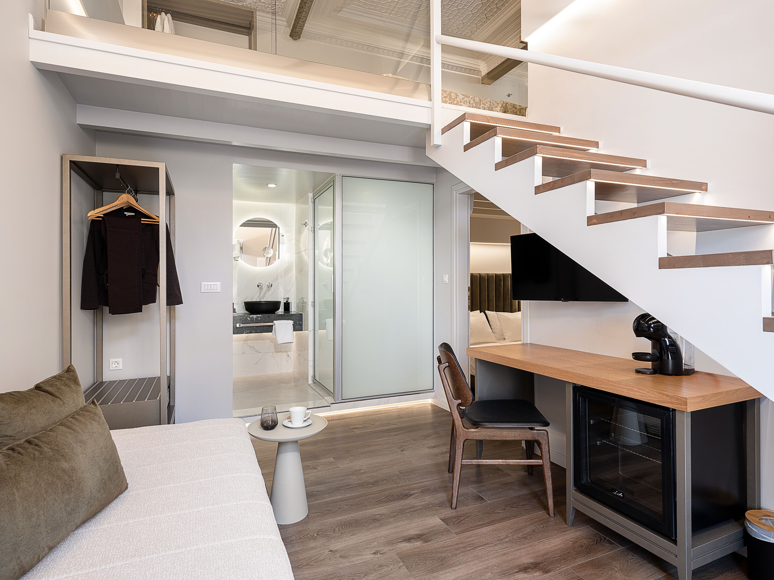 Ett modernt hotellrum med en soffa, ett litet bord, en stol, en kaffemaskin, en tv och en trappa.