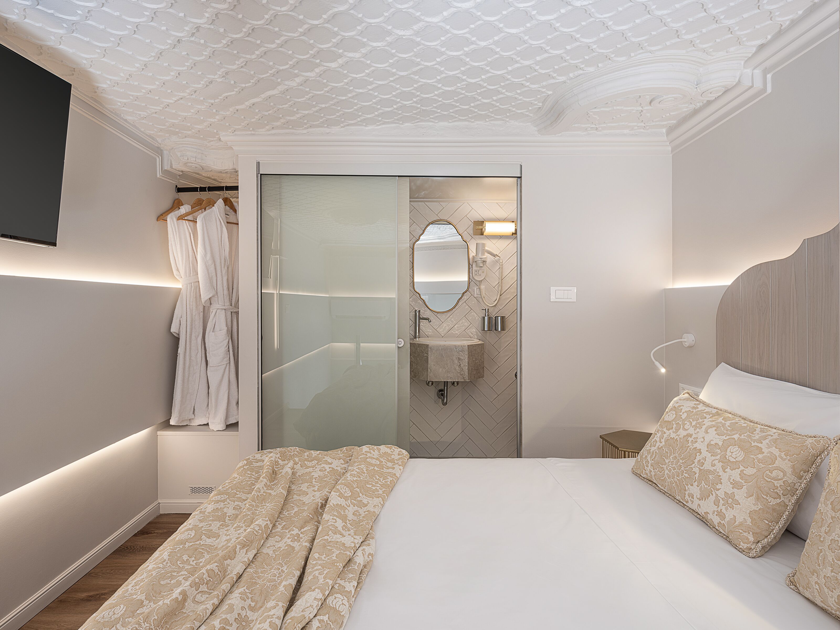 Une chambre moderne avec un grand lit, un dressing, une salle de bain avec un miroir et un lavabo, et un téléviseur fixé au mur.