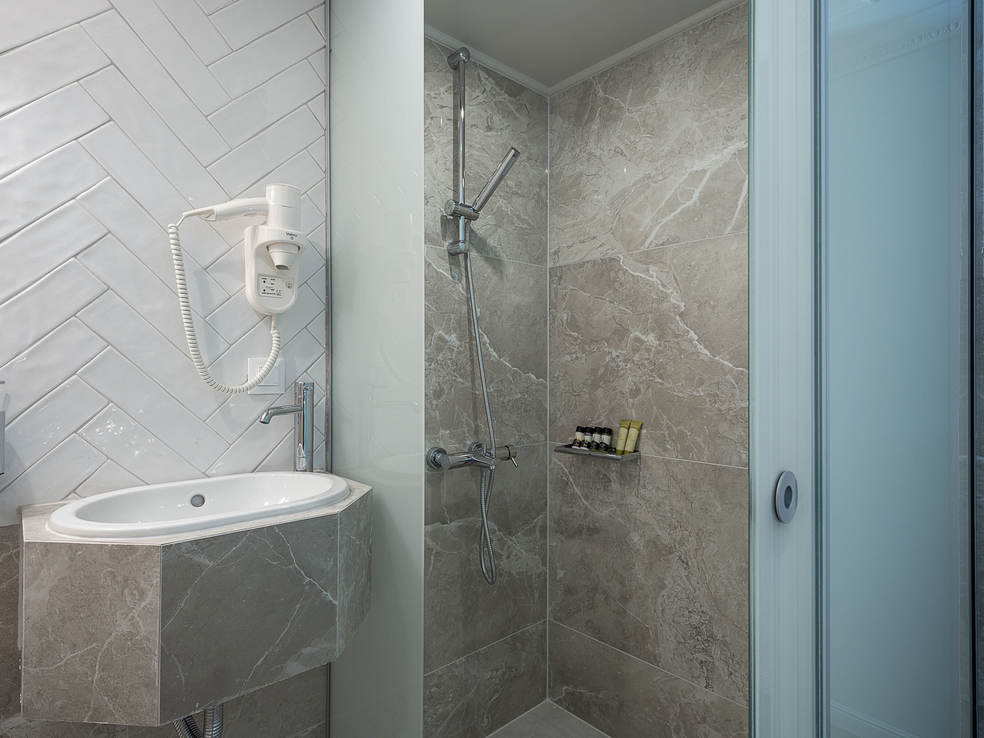 Un baño con lavamanos blanco, ducha con puerta de vidrio y zona de ducha con azulejos de mármol.