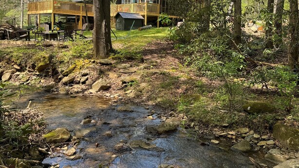 Mökki, jossa on terassi ja ulkona oleva oleskelualue, ja se sijaitsee metsän ja puron ympäröimänä.
