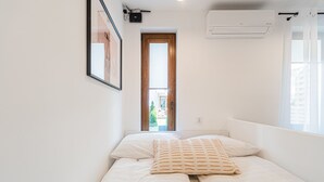 Un dormitorio pequeño y bien iluminado con cama, ventana con cortinas, unidad de aire acondicionado y puerta que da a un espacio exterior.