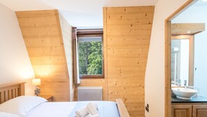 Ein Schlafzimmer mit Holzboden, einem Fenster mit Blick auf Bäume, einem Bett mit weißen Bettwäsche und einem Badezimmer mit Waschbecken und Spiegel.