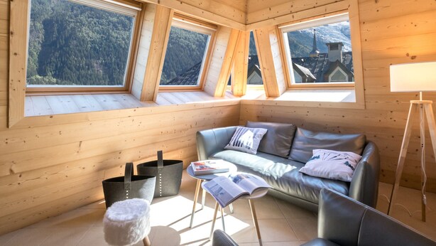 Ein gemütliches Zimmer mit Holzvertäfelung, einem Ledersofa, einem kleinen Tisch und einem Fenster mit Blick auf einen Wald und eine Kirche.
