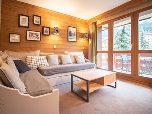 A cozy room with a wooden wall, a grey sofa, a wooden coffee table, and a large window with a view of trees.