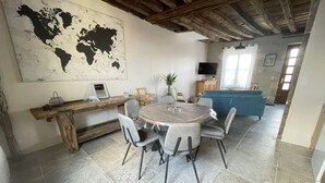 Een eetgedeelte met een ronde tafel, grijze stoelen, een houten bank, een wereldkaart aan de muur, een televisie en een blauwe bank op de achtergrond.