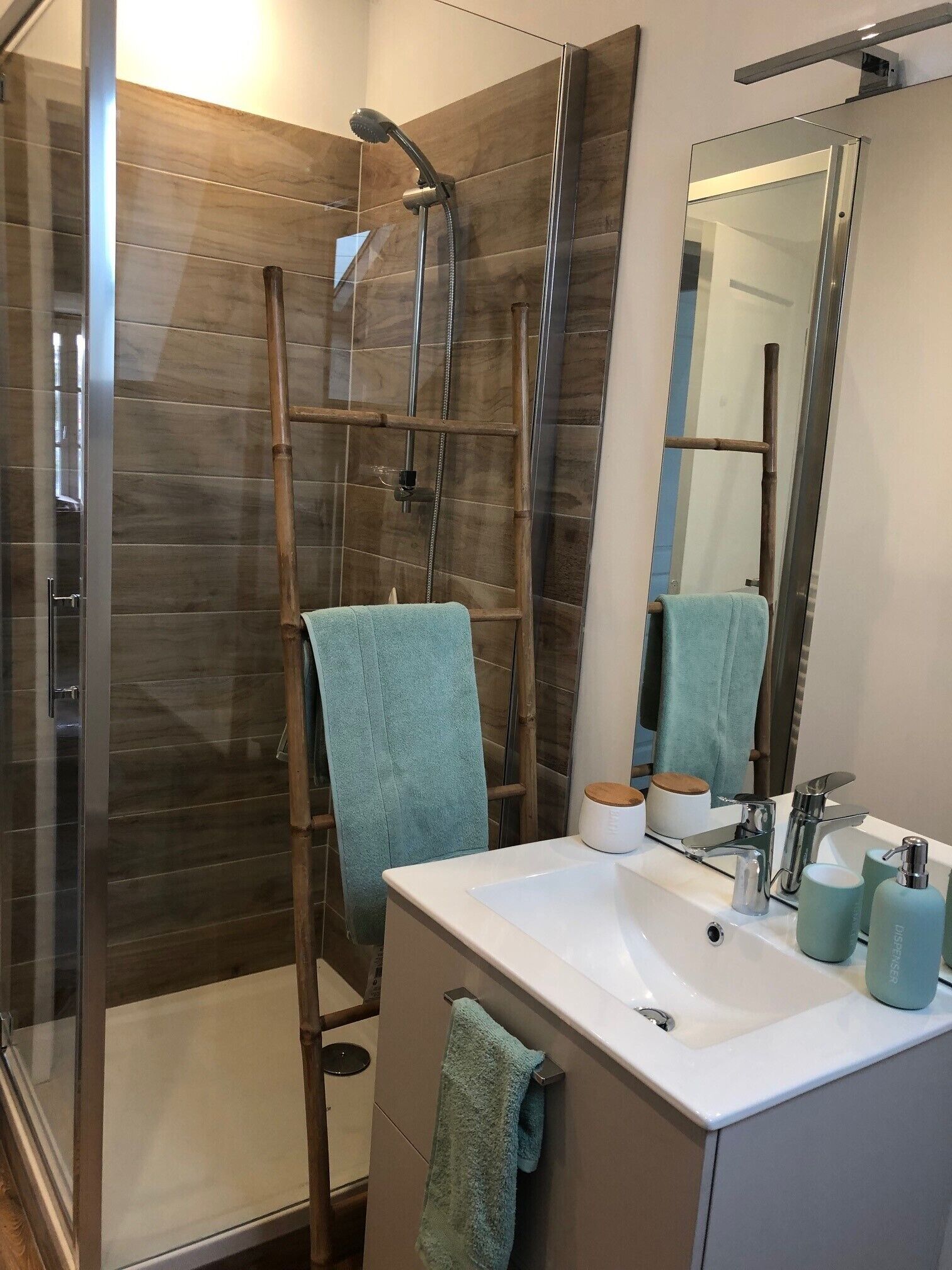 Une salle de bain moderne avec une douche à parois de verre, une échelle en bois, un lavabo blanc avec un miroir et un porte-serviettes.