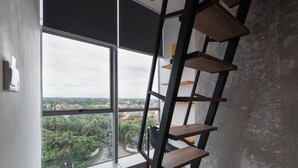 A room with a large window, a wooden ladder, and a view of a cityscape and greenery. - Cove Hillcrest (Curug)