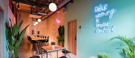 A modern interior with a neon sign stating "Relax nothing is under control", a wooden dining table, chairs, and potted plants.