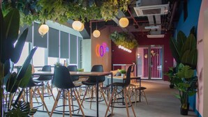 A modern office space with multiple tables and chairs, indoor plants, and a neon bicycle sign. - Cove Hillcrest (Curug)
