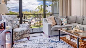 Una sala de estar con sofá, sillón y mesa de centro, un balcón con vistas a palmeras y montañas, y un ventanal con cortinas.