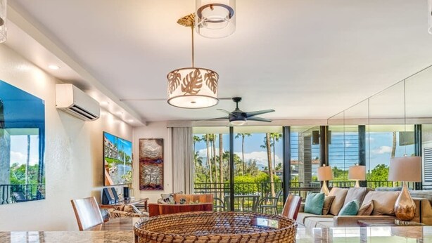 A spacious living area with a large round table, comfortable seating, and large windows offering a view of palm trees and a building. - Stunning Deluxe Ocean View W/Lanai-Kamaole Sands (Kihei)