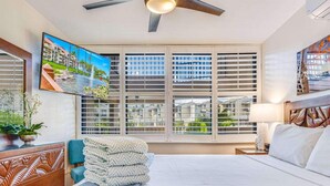 A hotel room with a bed, a ceiling fan, a window with blinds, a painting, and a nightstand with a lamp. - Stunning Deluxe Ocean View W/Lanai-Kamaole Sands (Kihei)
