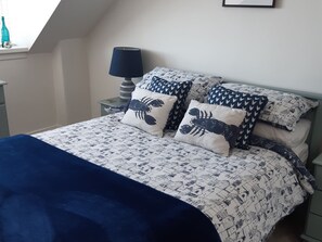 Una cama cuidadosamente hecha con una colcha de diseño azul y blanco, dos almohadas con motivos de cangrejo, una lámpara azul oscuro en una mesita de noche y un cuadro en la pared.