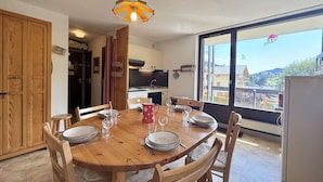 A dining area with a wooden table set for four, a kitchen in the background, and a large window offering a view of the outside. - 3-room apartment in Flumet - Parking - 6 people (Flumet)