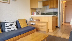 A compact living space with a sofa, a small wooden table, and a kitchenette area. - Cozy Apartment Near Slopes with Balconies (Montvalezan-La Rosière)