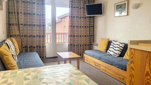A room with a sofa, a small table, a TV mounted on the wall, and a window with curtains. - Cozy Apartment Near Slopes with Balconies (Montvalezan-La Rosière)