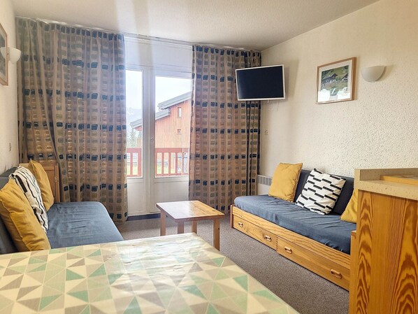 A room with a sofa, a small table, a TV mounted on the wall, and a window with curtains. - Cozy Apartment Near Slopes with Balconies (Montvalezan-La Rosière)