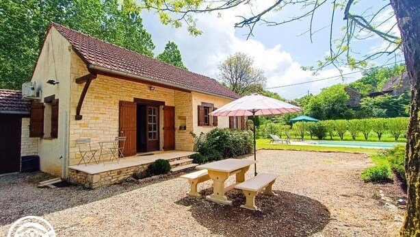 A stone house with a tiled roof, a wooden door, and a small porch. There is a picnic table with a pink umbrella in front. - La Petite Fagette (Frayssinet-le-Gélat)