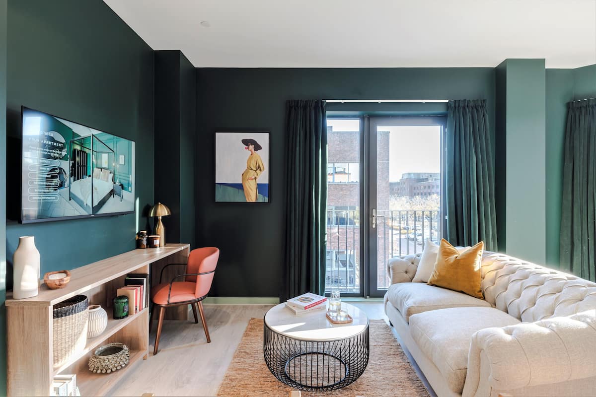 Un salon moderne avec un canapé, une table basse ronde et un bureau avec une chaise. Une grande fenêtre, ornée de rideaux, offre une vue sur les bâtiments environnants.