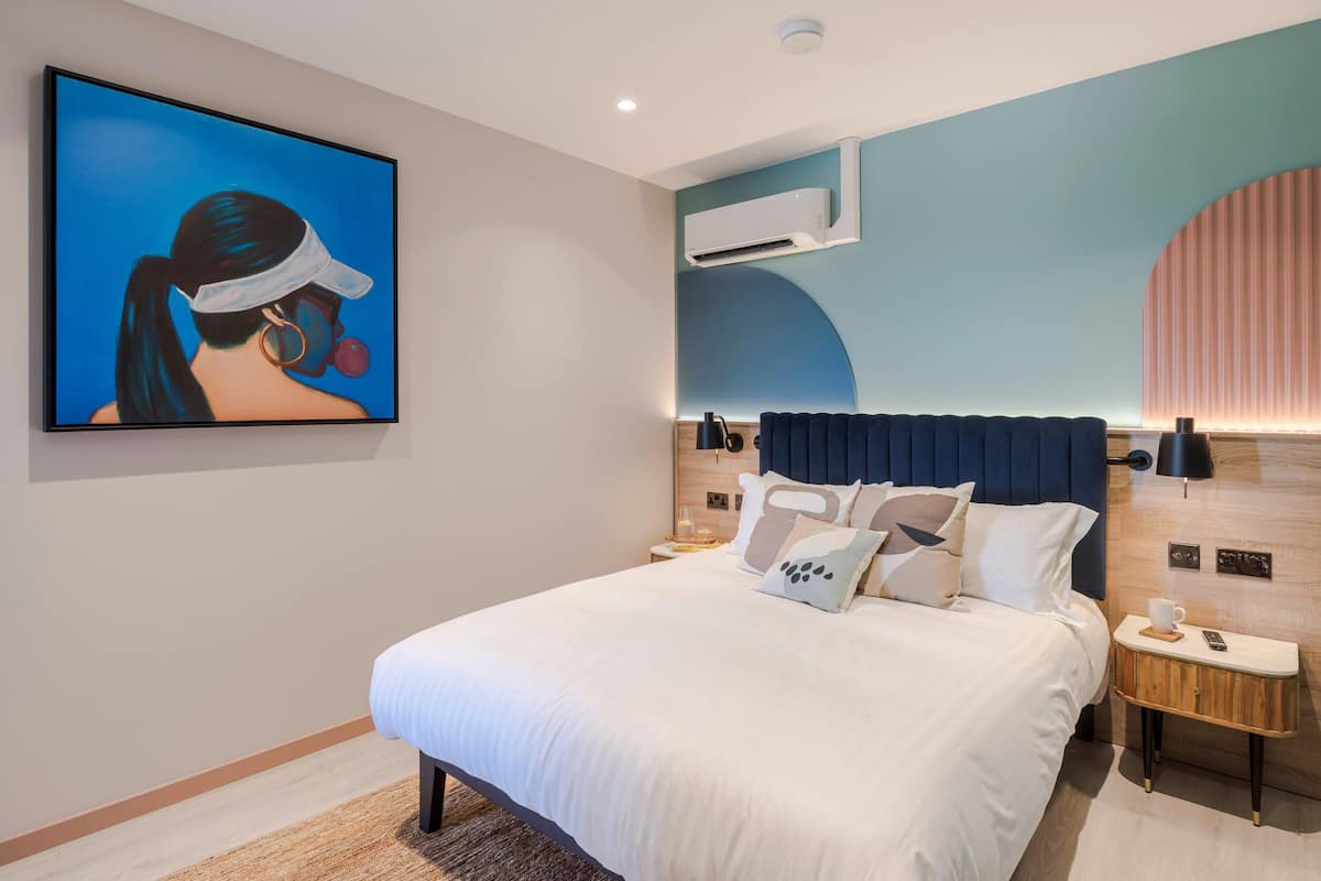 Une chambre d’hôtel moderne avec un grand lit, un climatiseur fixé au mur, une table de chevet avec une lampe et une œuvre d’art encadrée accrochée au mur.