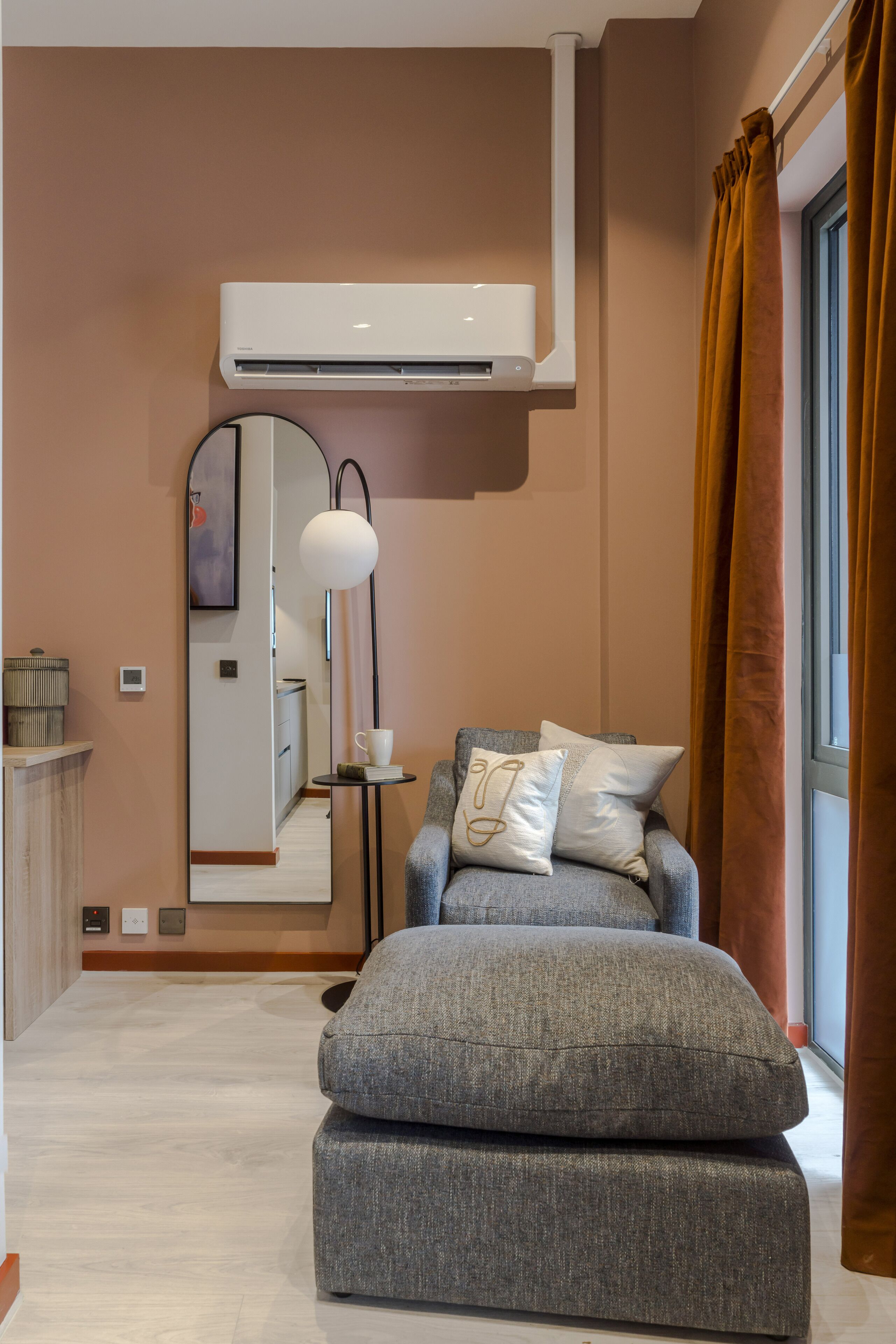 A modern living room with a grey sofa, a large mirror, and an air conditioning unit mounted on the wall.