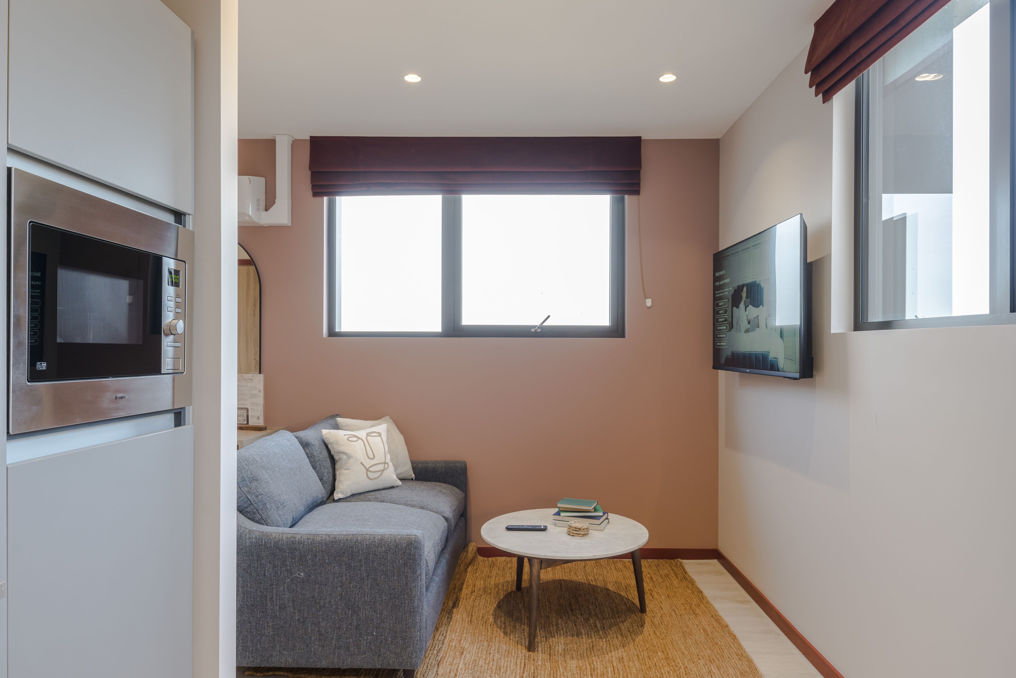 A compact living space with a grey sofa, a small round table, a mounted TV, and a window with blinds.