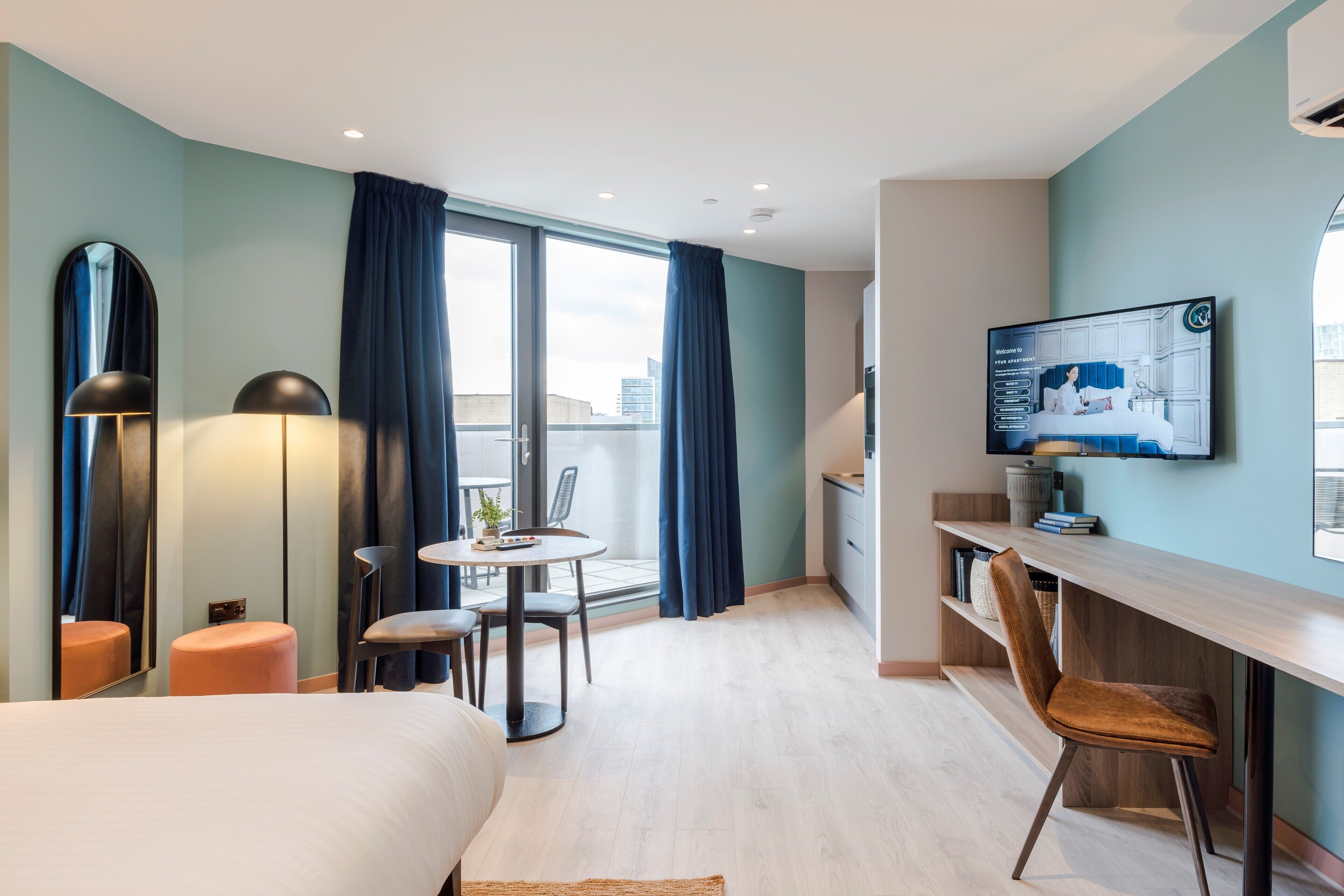 A modern hotel room with a large bed, a desk with a chair, a small table with chairs, a TV mounted on the wall, and a large window with curtains.