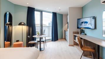 A modern hotel room with a large bed, a desk with a chair, a small table with chairs, a TV mounted on the wall, and a large window with curtains.