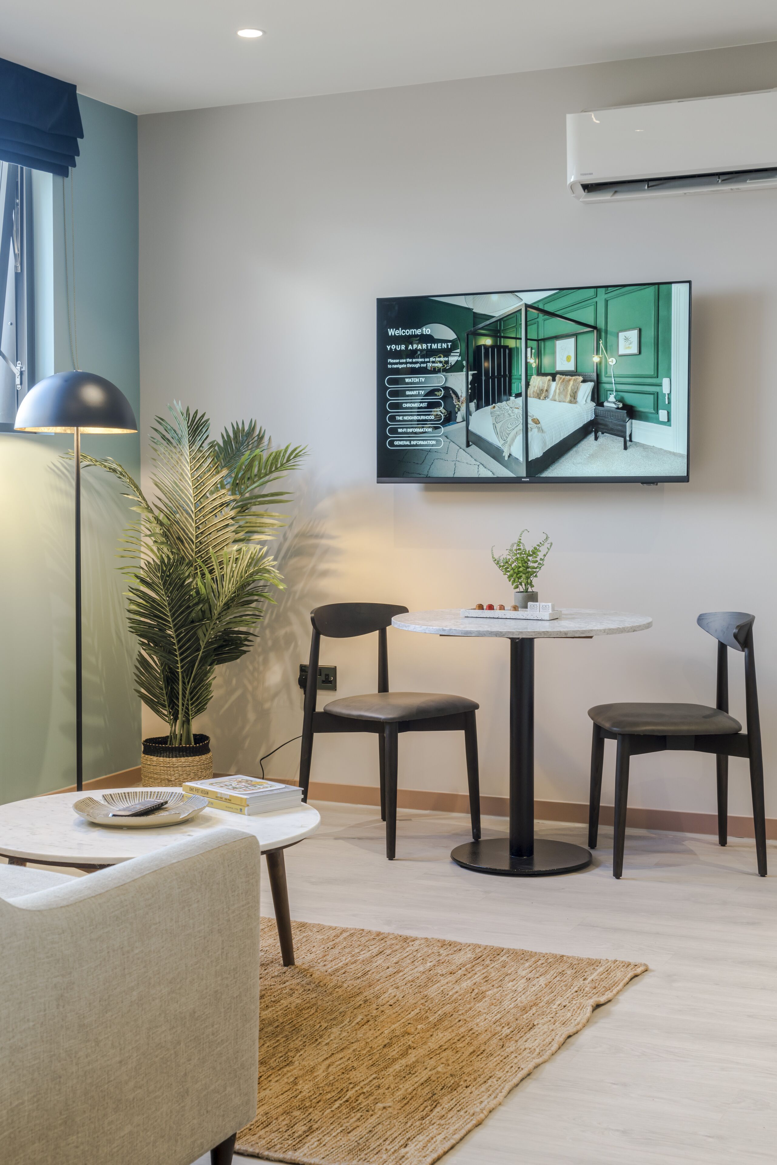 A modern hotel room with a large flat-screen TV displaying a welcome message, a round dining table with chairs, a potted plant, and a floor lamp.
