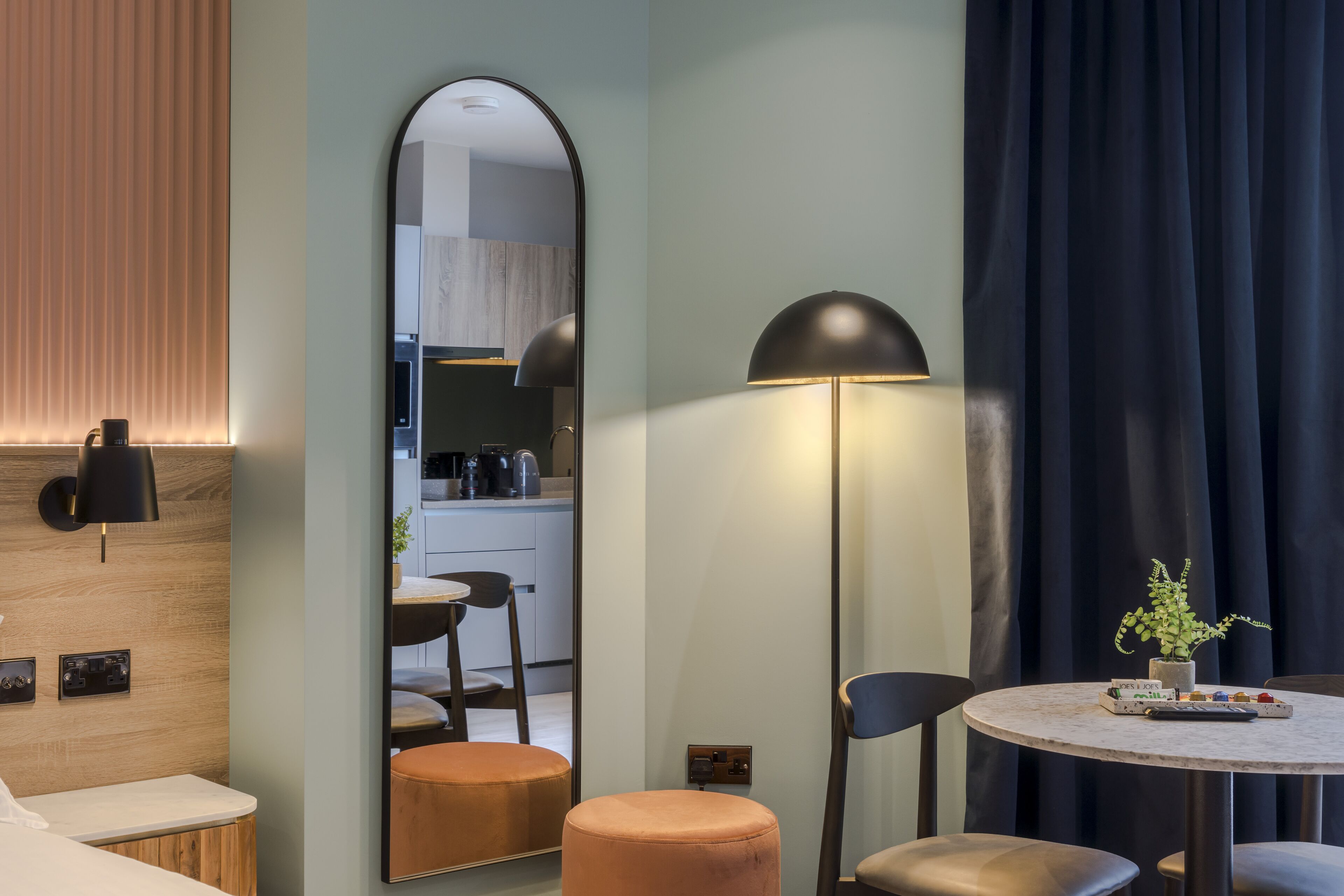 Ein modernes Hotelzimmer mit einer Wand in Holzoptik, einem runden Tisch mit einem Topfpflanzen und einem Spiegel, der den Küchenbereich reflektiert.