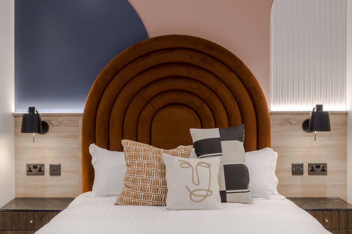 Une chambre à coucher moderne avec une tête de lit circulaire originale, un lit bien fait agrémenté d’oreillers à motifs et un éclairage mural.